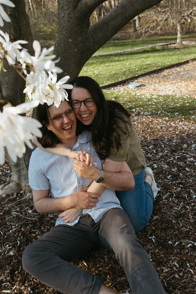 Couple sits under blossoming tree and laughs during spring engagement photos in Arnold Arboretum