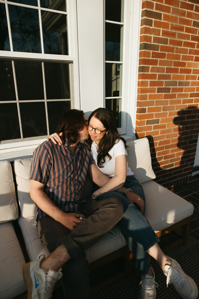 Couple sits on porch during golden hour and drinks wine during casual engagement session