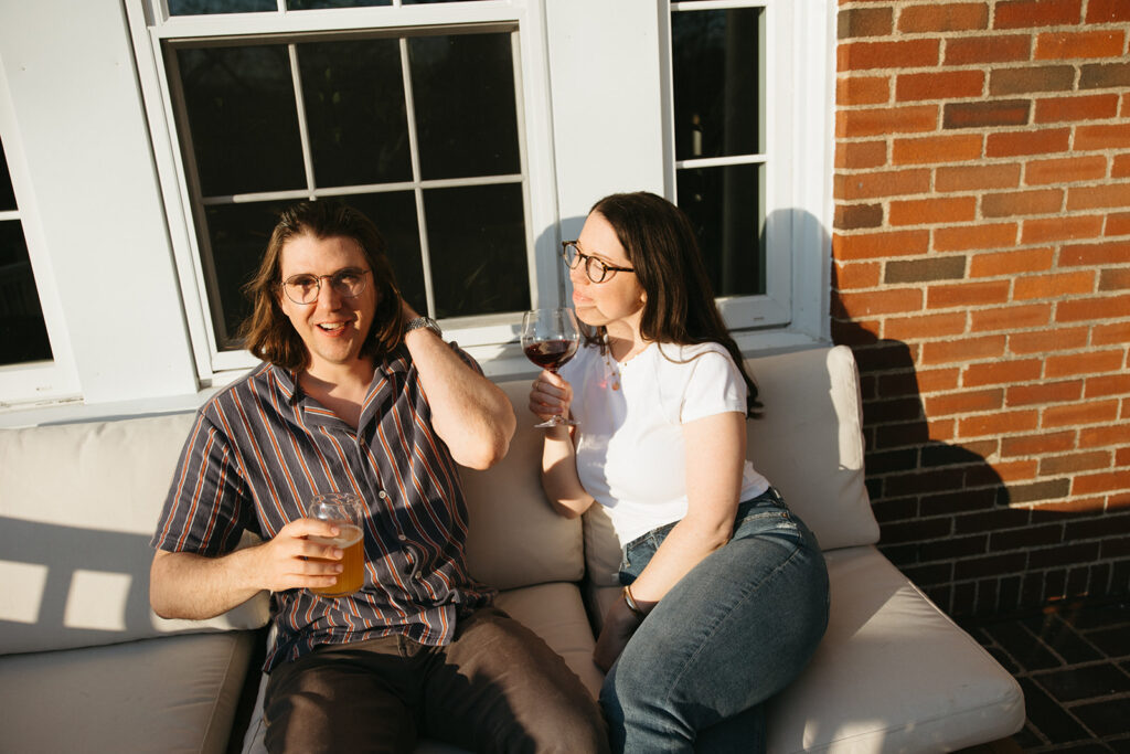 couple sits on porch with wine and beer during their laid back engagement session at home