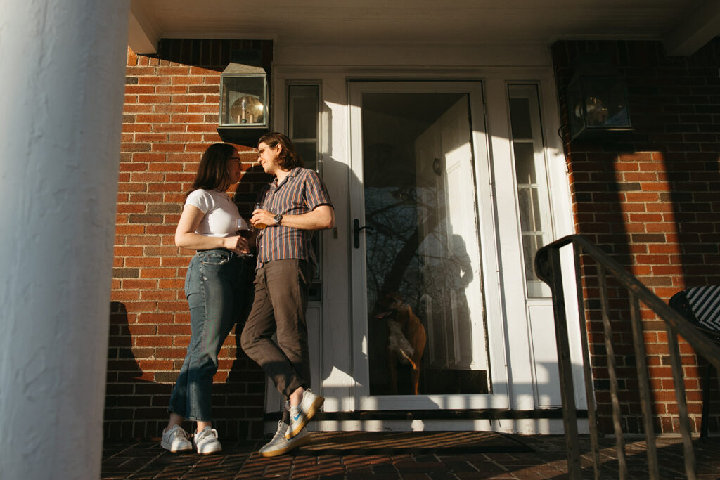 couple stands in front of their doorway on porch during engagement photos in boston