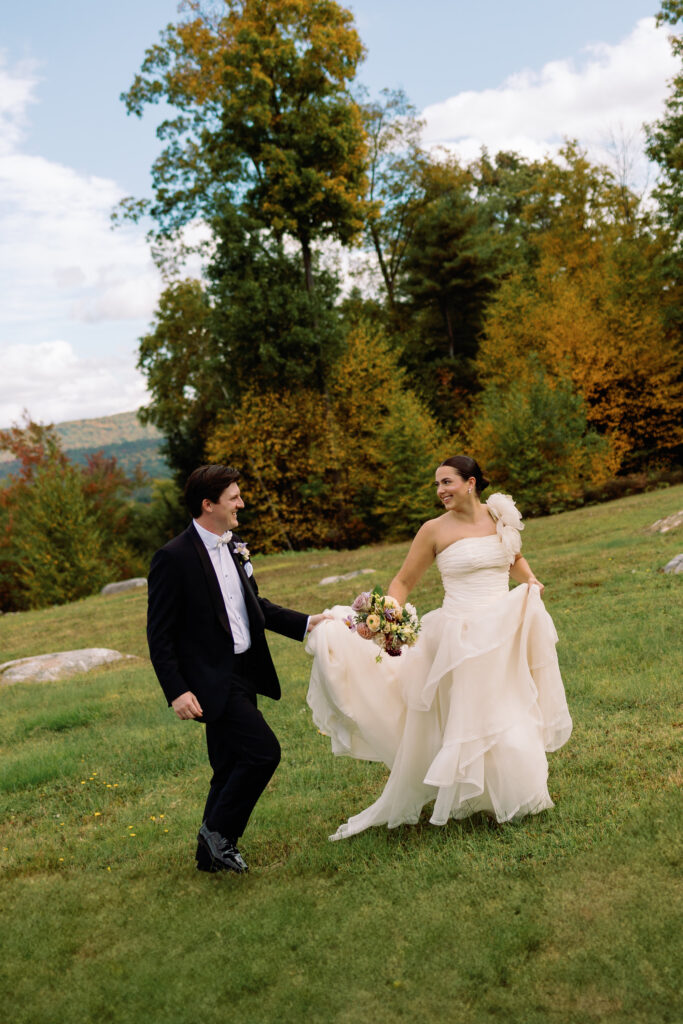 bride and groom run through field while groom holds her dress