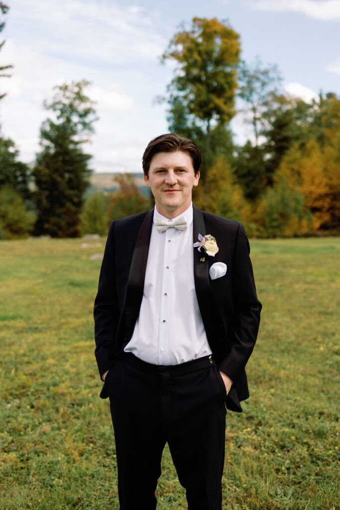 groom poses in field with fall foliage in the background