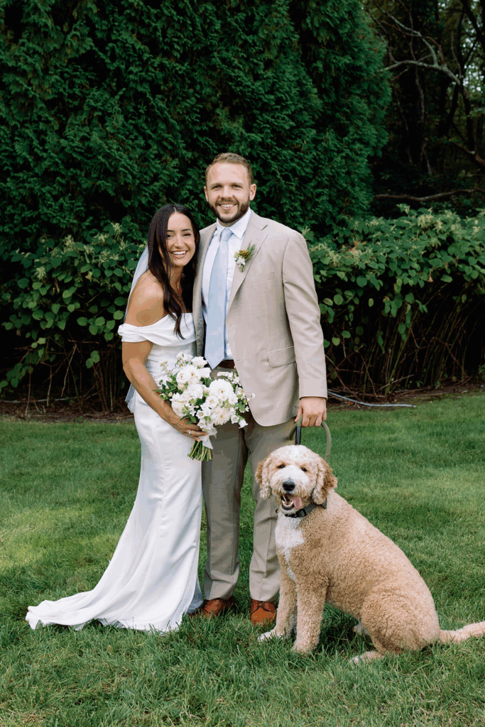 A groom and bride pose together smiling with their dog.