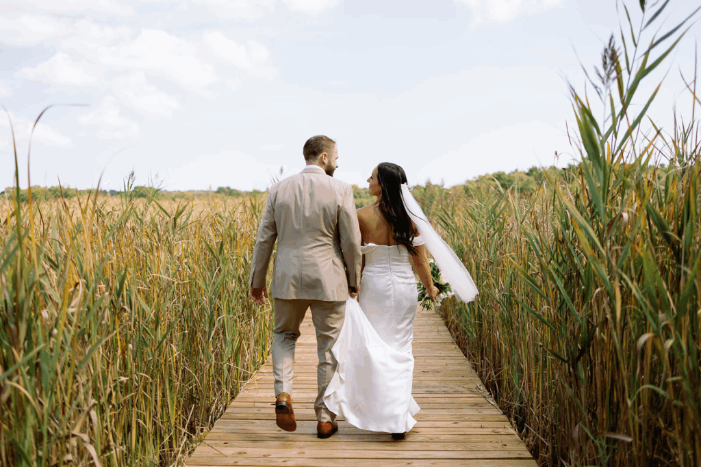 A bride and groom walk away together on a dock surrounded by reeds towards the horizon.