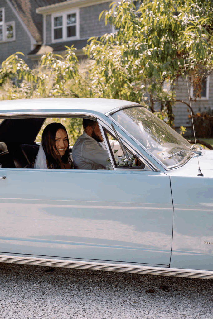 A groom drives his bride in a blue car as she smiles out the window.