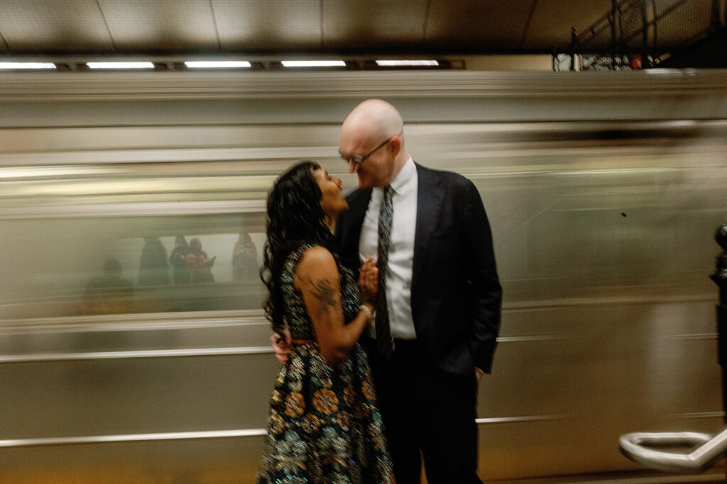 An engagement photo of a man and a woman holding and looking at each other as a train passes by in the background.